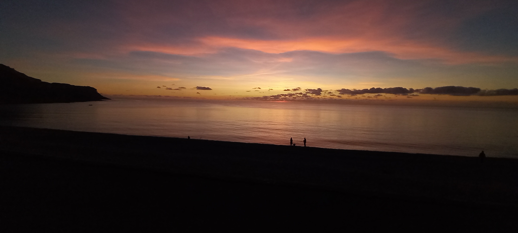 A pink and gold sunset over Chesil Beach with two figures at the water's edge